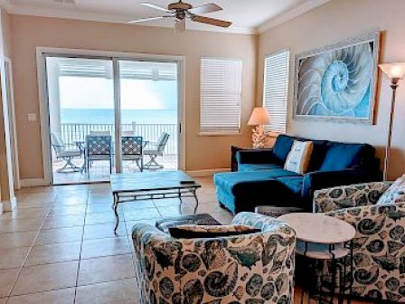 A cozy living room with ocean views, featuring patterned chairs, a blue sofa, a coffee table, and a painting of a nautilus shell.