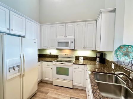 A kitchen with white cabinets, a fridge, oven, microwave, coffee maker, double sink, and a decorative plate on the counter.