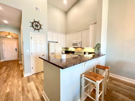 A kitchen with white cabinets, granite countertops, and bar stools. Wooden flooring, a ship wheel decor, and overhead lighting complete the look.