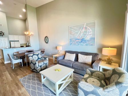 A cozy living room with a gray couch, patterned chairs, and a nautical-themed decor. There's a dining area and kitchen in the background.