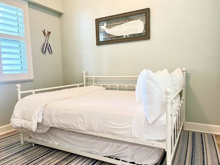 A minimalist bedroom features a white daybed, striped rug, paddle wall art, and a framed whale print for coastal decor.