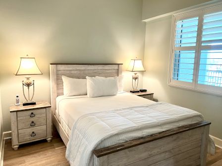 A cozy bedroom with a light wood bed, white bedding, two bedside tables with lamps, and a window with shutters.