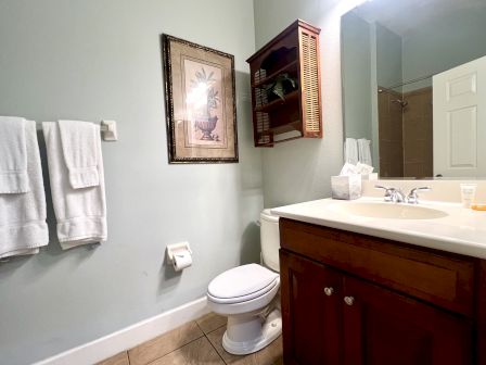 A bathroom featuring a toilet, sink with a wooden cabinet, mirror, towel rack with towels, a wall art piece, and a small wooden shelf.