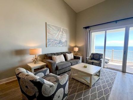 A cozy living room with ocean view features a sofa, armchairs, coffee table, and nautical-themed artwork, set by large windows leading outside.