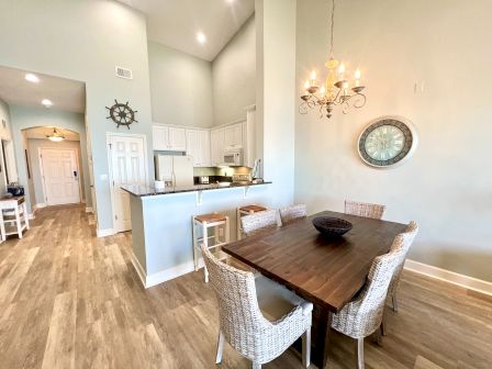 A dining area with a wooden table and six chairs, a kitchen bar with stools, chandelier, nautical decor, and a wall clock.