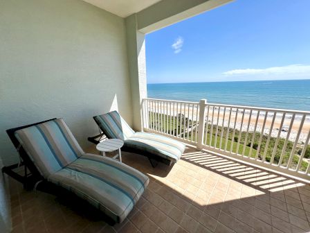 Two striped lounge chairs and a small table are on a balcony overlooking a sandy beach and clear blue ocean.