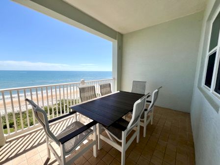 A balcony with a table and chairs, overlooking a beach and ocean under a clear blue sky.