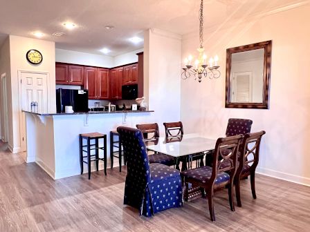 A dining area with a table, chairs, a chandelier, and a kitchen with wooden cabinets and bar stools. A clock is on the wall.