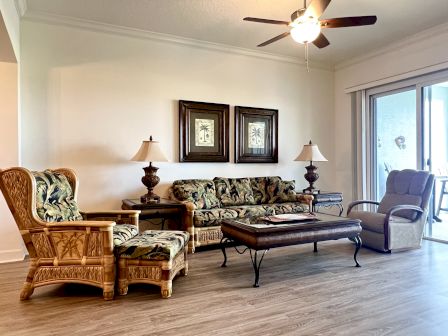 A living room with tropical-themed furniture, two lamps, a ceiling fan, sliding glass doors, and framed artwork on the wall.