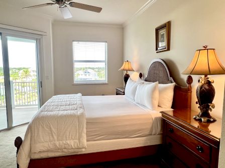 A cozy bedroom with a large bed, two ornate lamps, and a window offering a scenic view. A ceiling fan and artwork adorn the room.