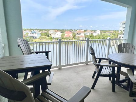 A balcony with wooden chairs and tables overlooks a serene lake view and colorful houses in the distance, under a clear blue sky.