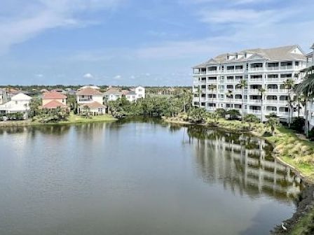 A serene lakeside scene with a large building and several houses in the background, under a clear blue sky.