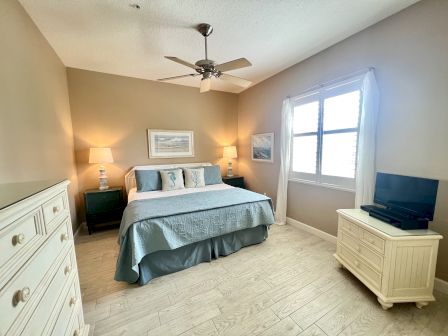 A cozy bedroom with a bed, lamps, dresser, TV, and window, decorated in neutral tones with blue accents.
