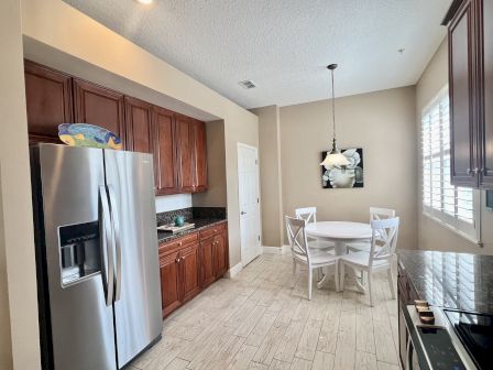 A kitchen with stainless steel fridge, wooden cabinets, round table with chairs, hanging light, and flower artwork on the wall.