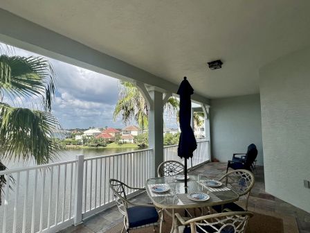 A covered patio with a glass table, chairs, and large umbrella overlooking a scenic waterfront with palm trees and houses in the distance.