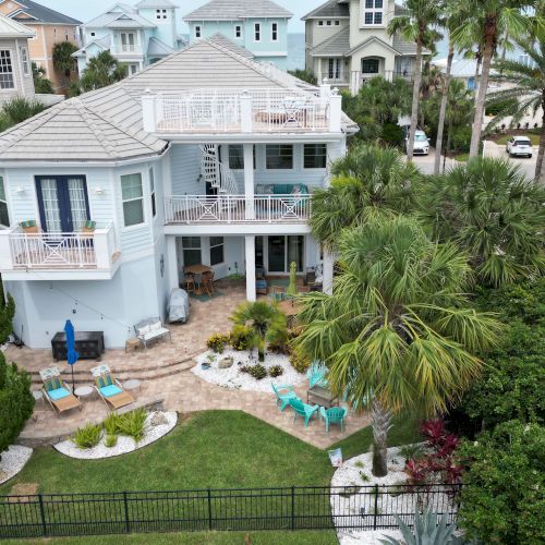 A large two-story house with multiple balconies, a landscaped yard, and outdoor seating surrounded by palm trees and greenery.
