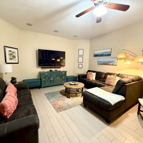 A cozy living room with sofas, a large TV, a coffee table, and decorative lamps. The walls feature framed pictures and a ceiling fan.