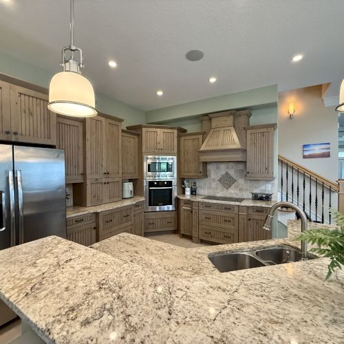 A modern kitchen with wooden cabinets, stainless steel appliances, granite countertops, pendant lights, and a plant.