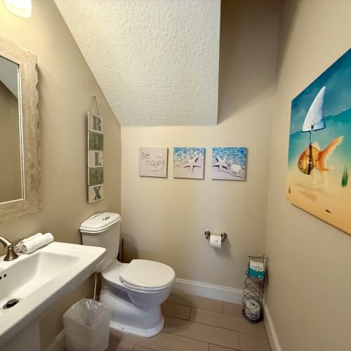 A small bathroom with a sink, toilet, mirror, and ocean-themed decor, including a fish artwork and signs saying "Relax" and "Be Happy."
