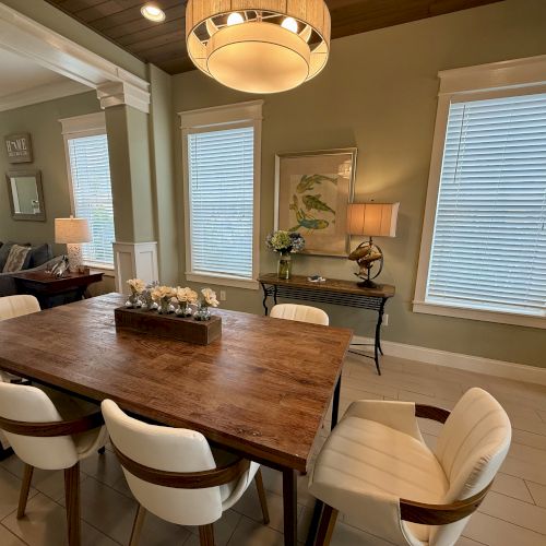 A dining area with a wooden table, cream chairs, and decorative plants, illuminated by a hanging light with a cozy living space nearby.
