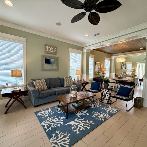 A cozy living room features a blue sofa, decorative pillows, tables, lamps, a blue rug with a coral pattern, and an open dining area.