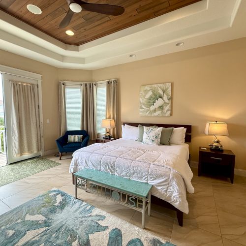 A cozy bedroom with a king-sized bed, decorative pillows, blue chair, ceiling fan, and a view of a balcony through sliding glass doors.