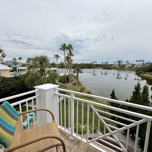 A balcony overlooking a serene lake with palm trees and houses in the background, featuring a wicker chair with a colorful cushion.