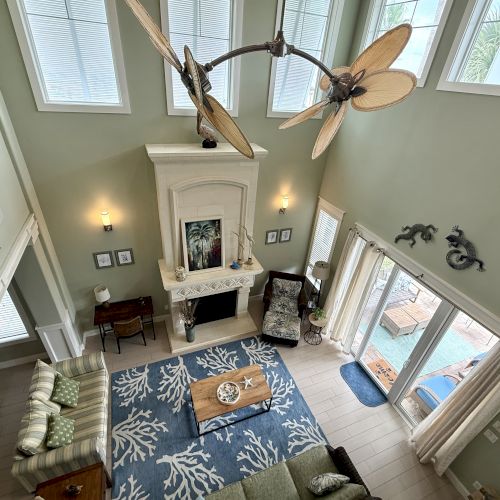 A spacious living room with high ceilings, a fan, fireplace, patterned rug, and large windows overlooking a pool area.
