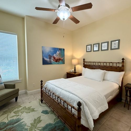 A cozy bedroom with a wooden bed, white bedding, chairs, art on the wall, and a ceiling fan above, warmly lit by lamps.