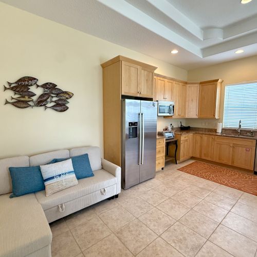 A cozy kitchen and living area with light wood cabinets, a sofa, cushions, fish wall art, and modern appliances on tiled flooring.