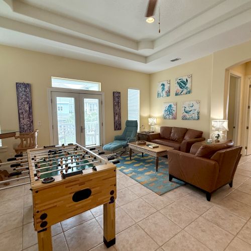 A living room with a foosball table, couch, armchair, and wall art; large windows provide natural light, and there's a glimpse of a hallway.