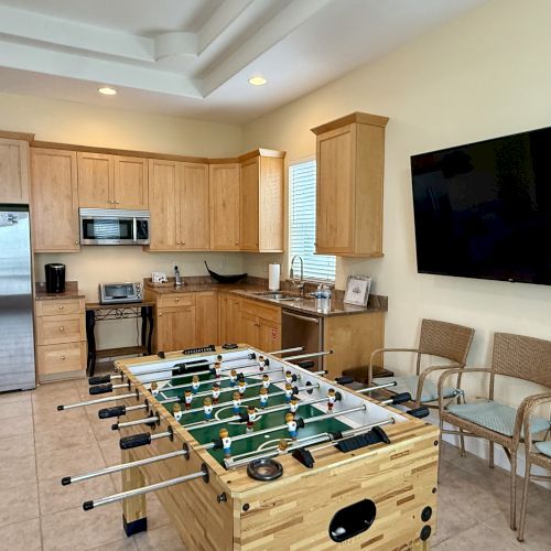 The image shows a kitchen area with a foosball table, a large TV, and chairs. The kitchen has wooden cabinets and modern appliances.
