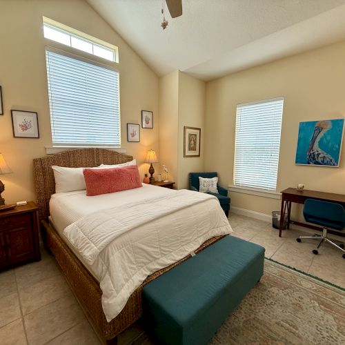 A cozy bedroom with a bed, side tables, lamps, art on the walls, a desk with a chair, and a blue ottoman at the foot of the bed.