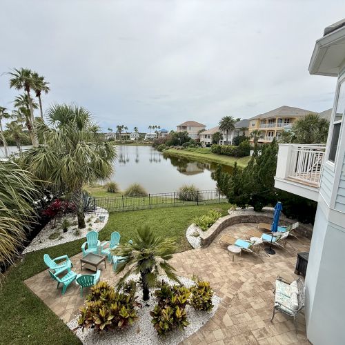 The image shows a lakefront backyard with patio seating, palm trees, and a view of nearby houses and greenery under a cloudy sky.