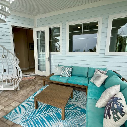 Outdoor patio with teal L-shaped sofa, tropical cushions, wooden table, spiral staircase, and blue walls, creating a cozy, inviting space.