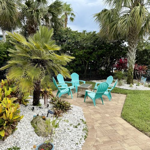 A backyard patio features turquoise chairs around a table, surrounded by palm trees, plants, white pebbles, and a paved path.