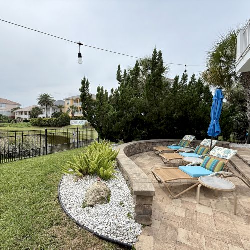 A patio with lounge chairs, umbrella, and string lights overlooks a grassy area and pond, surrounded by trees and neighboring houses.