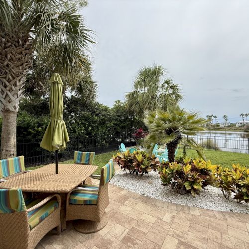 A patio with wicker furniture and a green umbrella overlooks a garden and a pond, surrounded by palm trees and lush greenery.