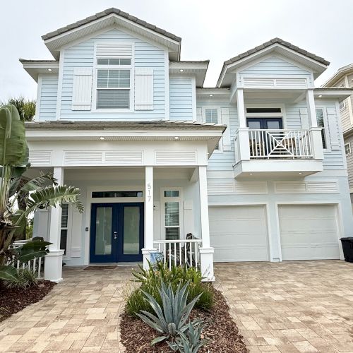 A modern two-story house with a pale blue exterior, white shutters, and a two-car garage is shown with tropical plants in the foreground.