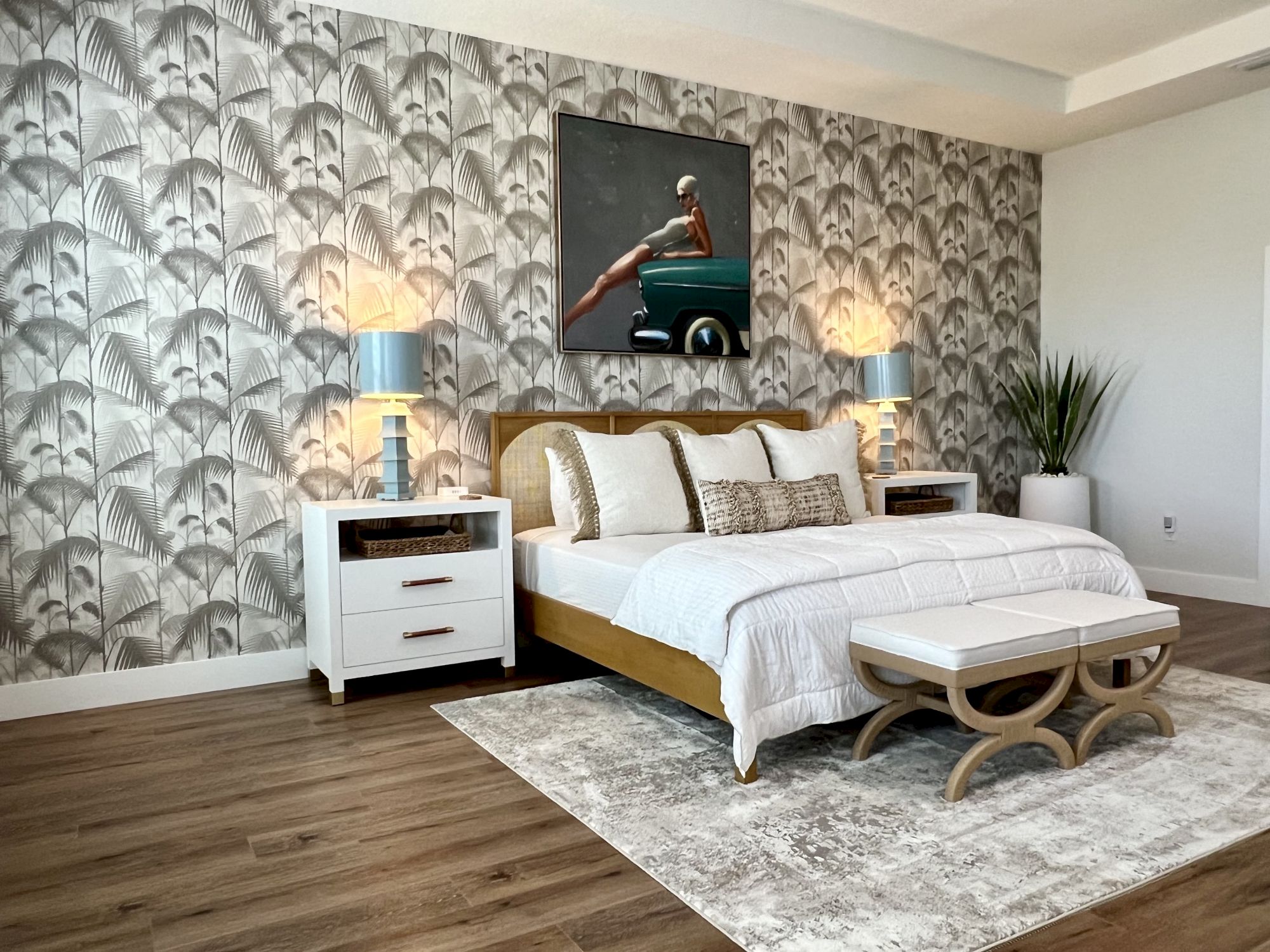 A stylish bedroom with palm leaf wallpaper, a bed with white linens, side tables with lamps, an artwork above, and a plant in the corner.