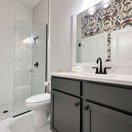 A modern bathroom features a glass shower, toilet, and dark vanity with a wide mirror, accented by a vibrant, patterned wall.