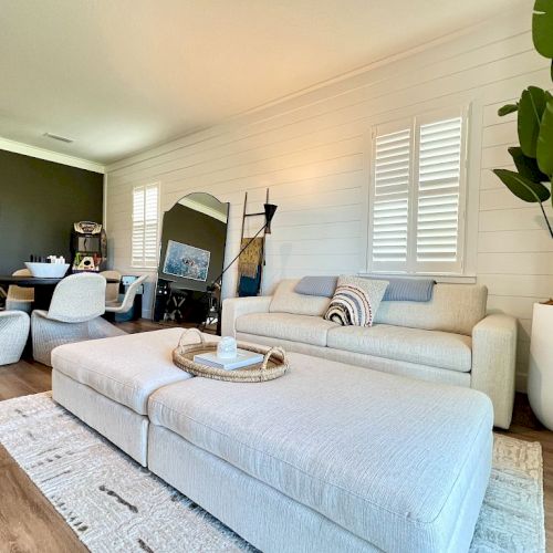 A modern living room with a large white sofa, a big ottoman, a plant, a dining table with chairs, and a wall with shutters and decor.