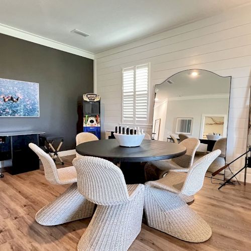 A cozy room with a round table, six woven chairs, large mirror, decorative ladder, artwork, and a dark accent wall.