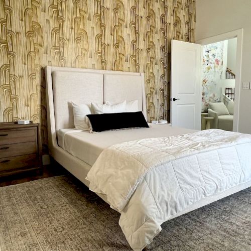 A bedroom with a patterned wallpaper, bed, wooden nightstand, lamp, and a round mirror on the wall is shown.