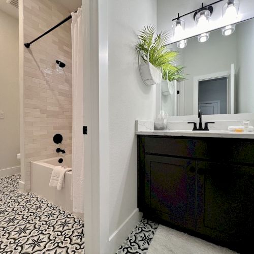 A modern bathroom with patterned floor, dark vanity, round mirror, and bathtub with curtain; decorated with potted plant on the wall.