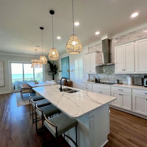 A modern kitchen with marble island, bar stools, pendant lights, white cabinets, and wood flooring; open layout leading to a bright area.