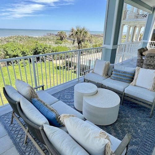 A coastal patio features white cushioned seating, round tables, and ocean views, with a lush garden and palm trees in the background.