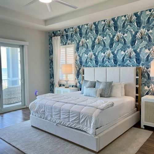 A bedroom with blue floral wallpaper, a bed, nightstands, lamps, and a view of the ocean through sliding glass doors is shown.