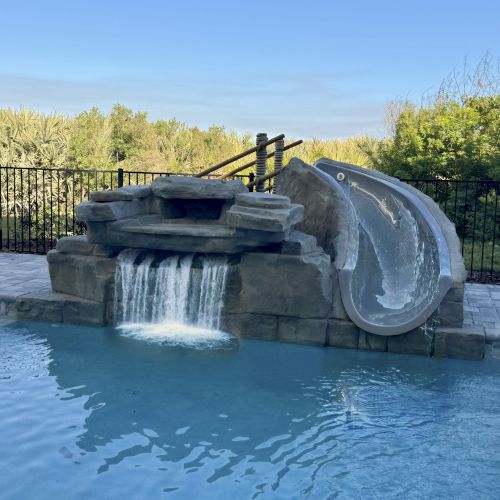 A swimming pool with a slide and waterfall feature, surrounded by a black fence and trees in the background.