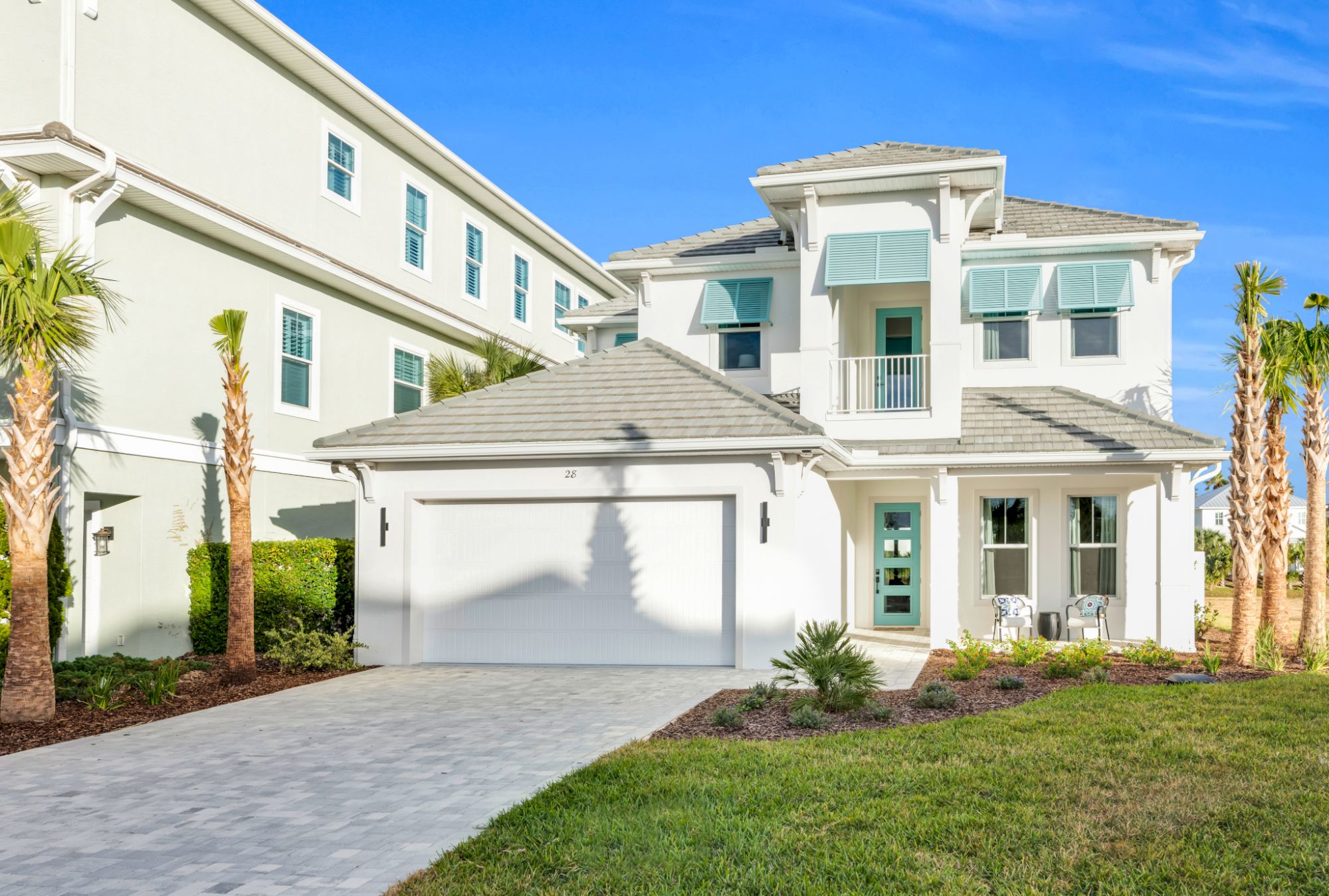 A modern two-story house with a garage, light-colored façade, green accents, a driveway, and manicured landscaping under a clear blue sky.